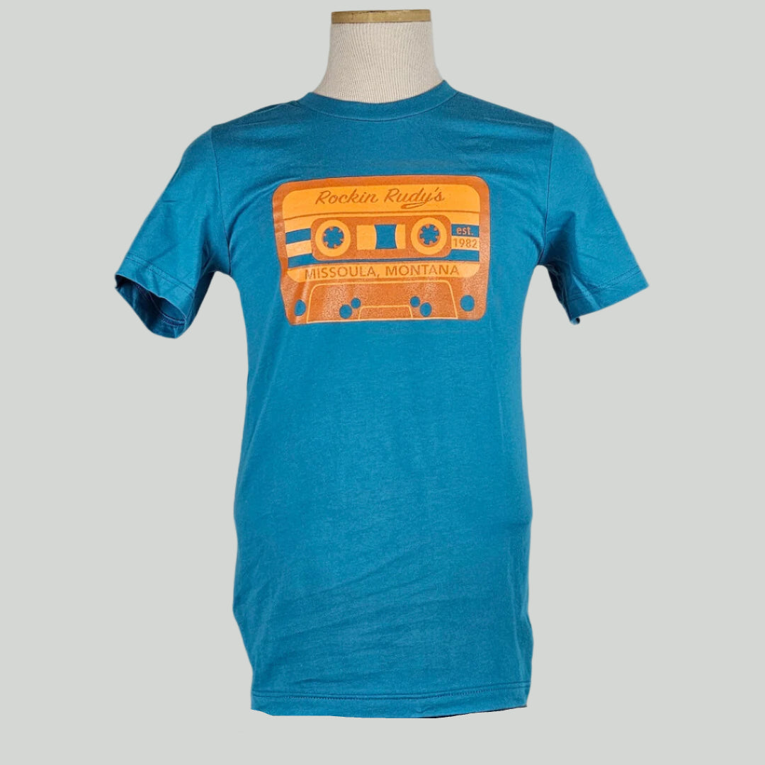 Blue shirt with blue & orange cassette tape across chest. Tape reads "Rockin Rudy's, est. 1982, Missoula, Montana."