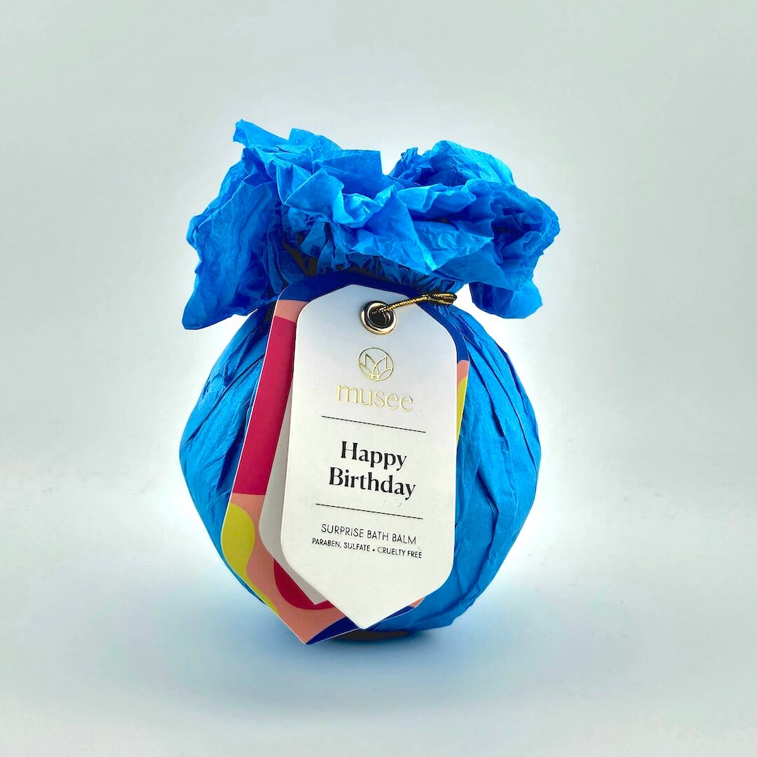 Bath bomb wrapped in blue tissue paper with a 'Happy Birthday' tag on a light gray background