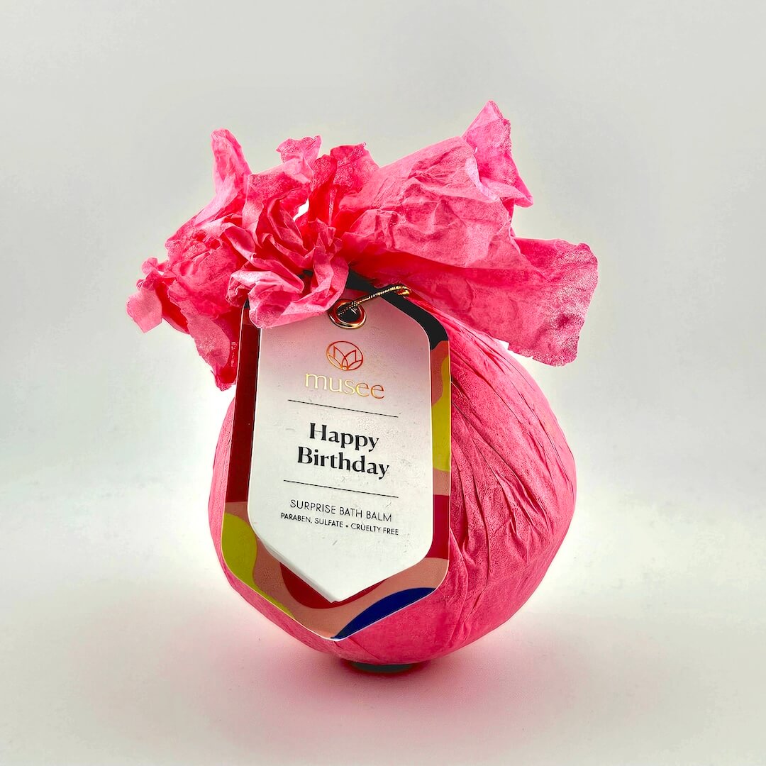 Pink wrapped bath bomb with a 'Happy Birthday' tag on a light gray background