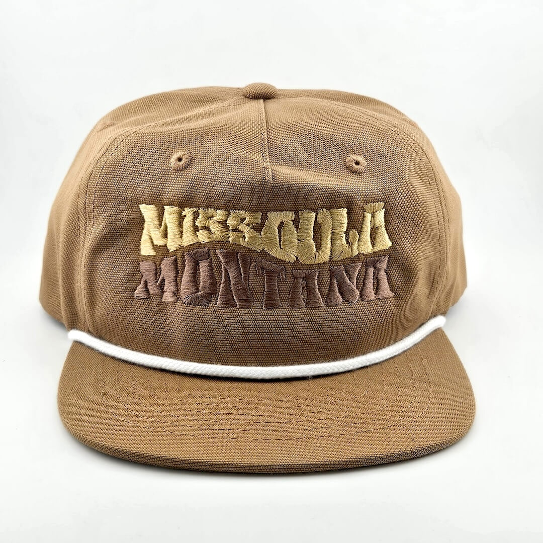 Brown canvas hat with "MISSOULA, MONTANA," in wavy block letters across face. White cord sitting on top of brim against face.