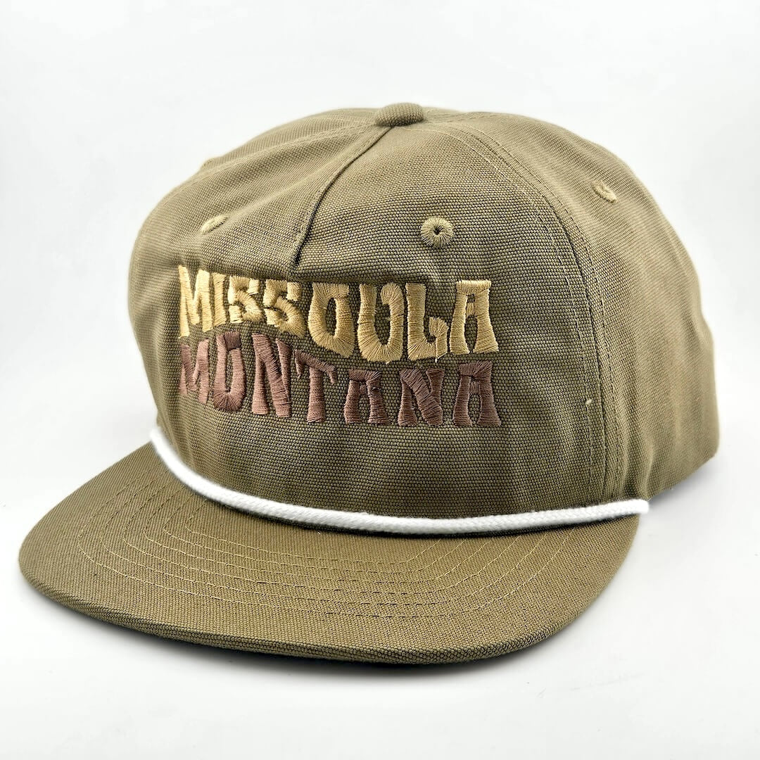 Green canvas hat with "MISSOULA, MONTANA," in wavy block letters across face. White cord sitting on top of brim against face.