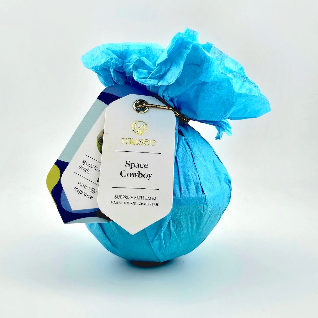 Blue bath bomb with a tag labeled 'Space Cowboy' on a light gray background