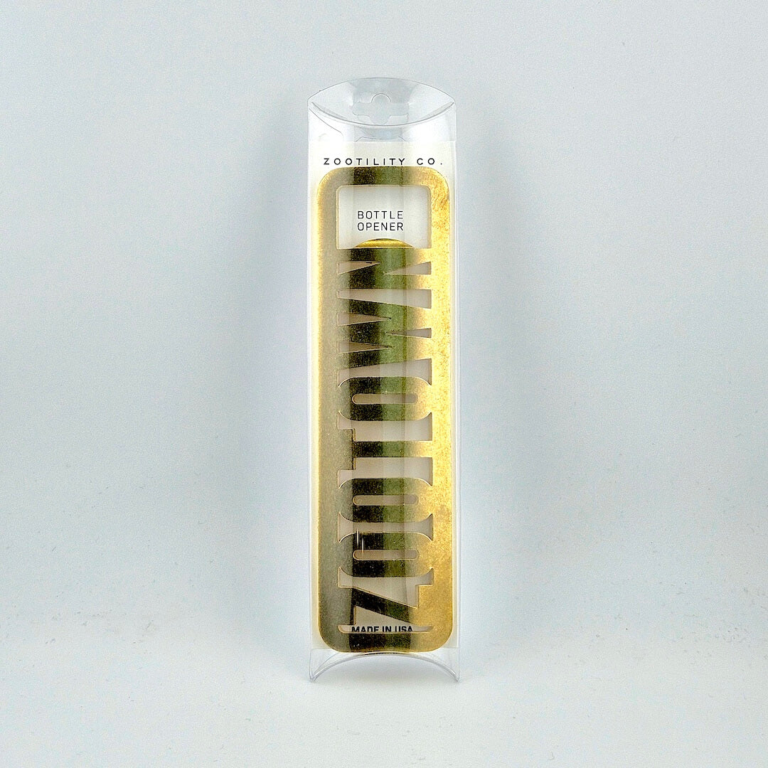 Brass bottle opener reading "ZOOTOWN" in packaging against white background.