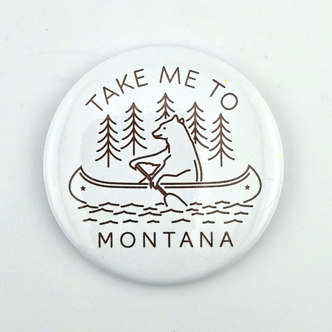 Button badge with bear in a canoe and text 'Take Me to Montana' on a white background