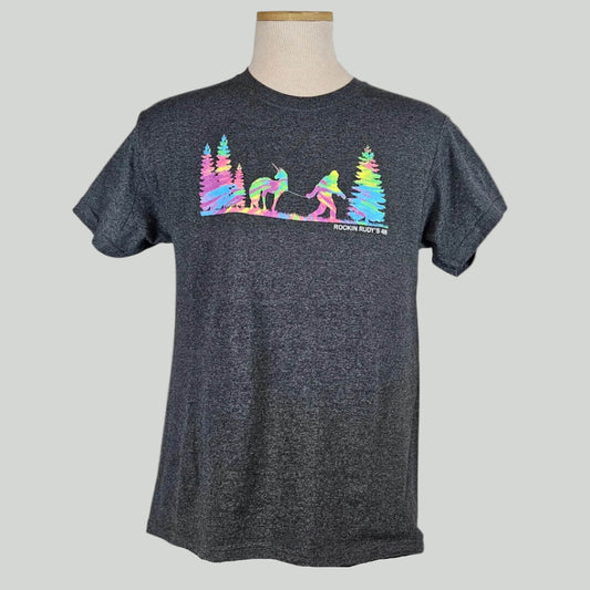 Dark heathered gray shirt with silhouette of trees either side of Bigfoot leading a unicorn on a leash in rainbow tie dye. 