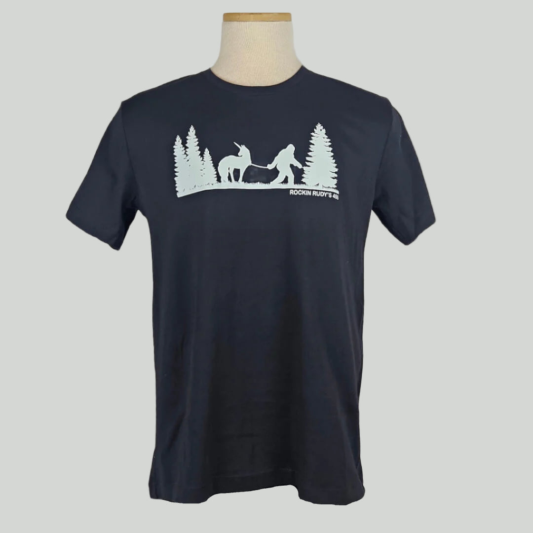 Black shirt with silhouette of trees either side of Bigfoot leading a unicorn on a leash in white print. 