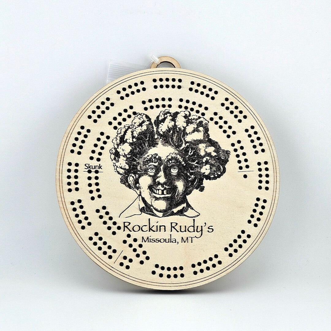 Circular wooden cribbage board with broccoli-haired man's face in center above "Rockin Rudy's, Missoula, MT"