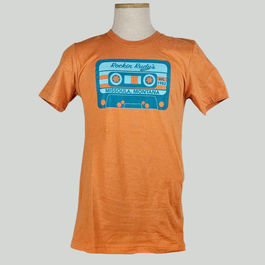 Orange shirt with blue & orange cassette tape across chest. Tape reads "Rockin Rudy's, est. 1982, Missoula, Montana."