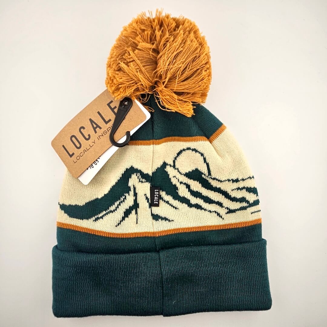 Knitted beanie with mountain design and orange pom-pom, attached to a 'Locale' tag on a light beige background.