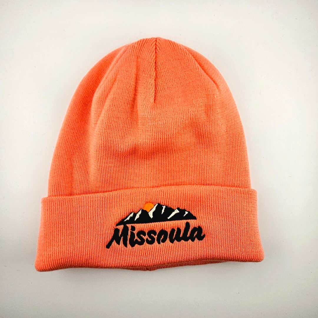 Salmon pink beanie with "Missoula" embroidered across cuff under mountain design.