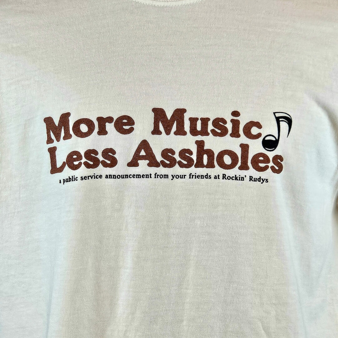 beige t-shirt with brown text 'More Music Less Assholes' and a black music note graphic on a beige background.