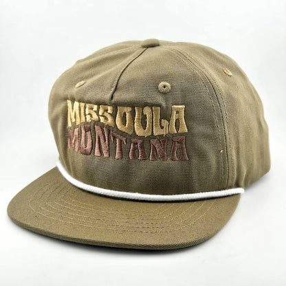 Green canvas hat with "MISSOULA, MONTANA," in wavy block letters across face. White cord sitting on top of brim against face. 