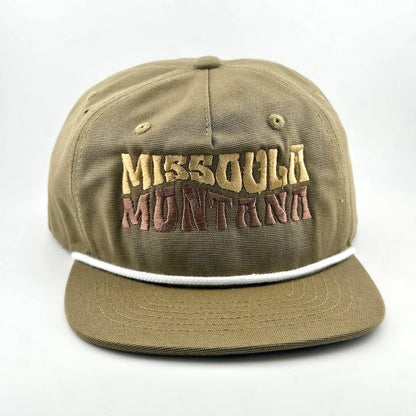 Green canvas hat with "MISSOULA, MONTANA," in wavy block letters across face. White cord sitting on top of brim against face. 