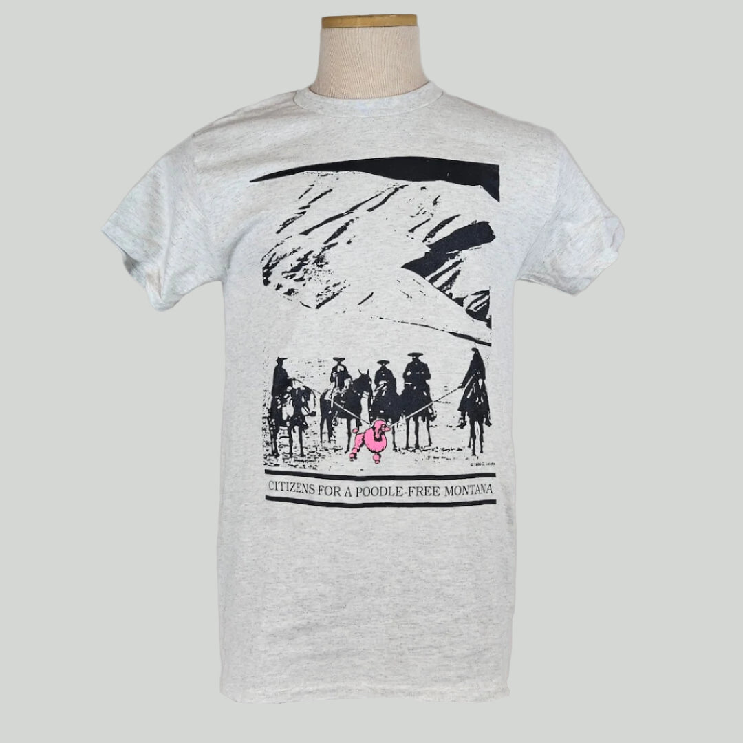 Gray shirt with mountain landscape behind group of mounted cowboys herding pink poodle with lassos. Caption under image. 