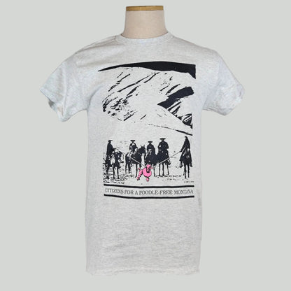 Gray shirt with mountain landscape behind group of mounted cowboys herding pink poodle with lassos. Caption under image. 