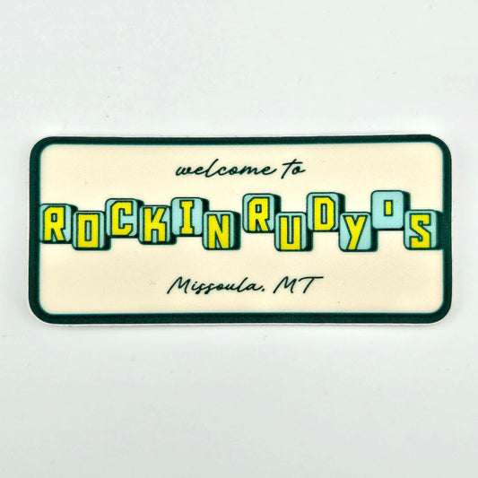 Rectangular sign with 'welcome to Rockin' Rudy's, Missoula, MT' text on a beige background