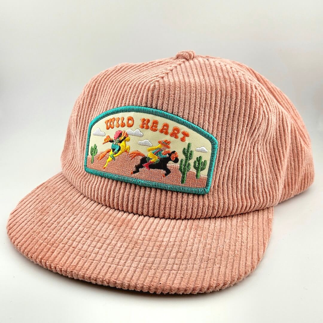 Pink corduroy cap with 'Wild Heart' patch featuring a cowboy and cowgirl design on a light gray background.