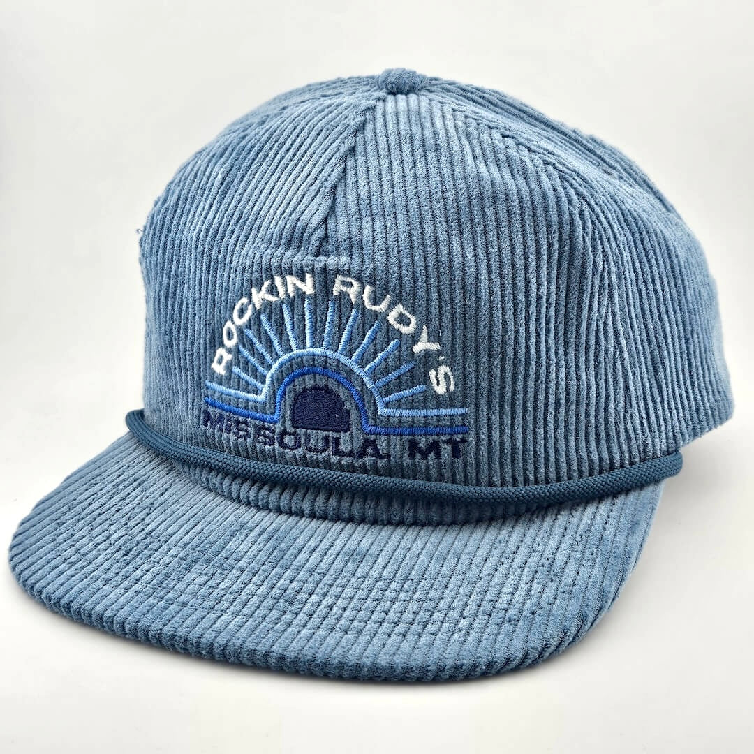 Blue corduroy billed hat with "ROCKIN RUDY'S", radiating from sunburst, "MISSOULA, MONTANA" along horizon bottom.