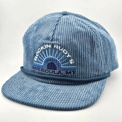 Blue corduroy billed hat with "ROCKIN RUDY'S", radiating from sunburst, "MISSOULA, MONTANA" along horizon bottom.
