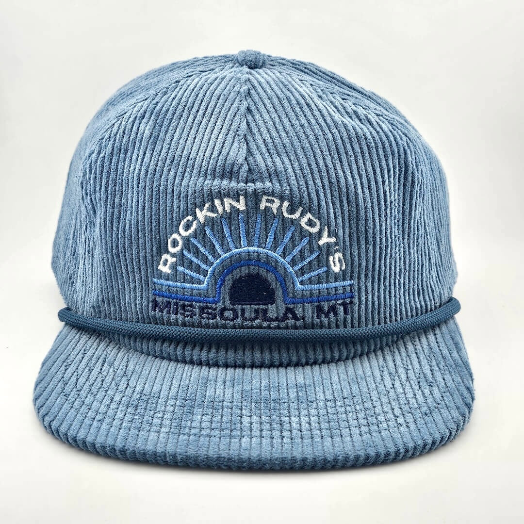 Blue corduroy billed hat with "ROCKIN RUDY'S", radiating from sunburst, "MISSOULA, MONTANA" along horizon bottom.