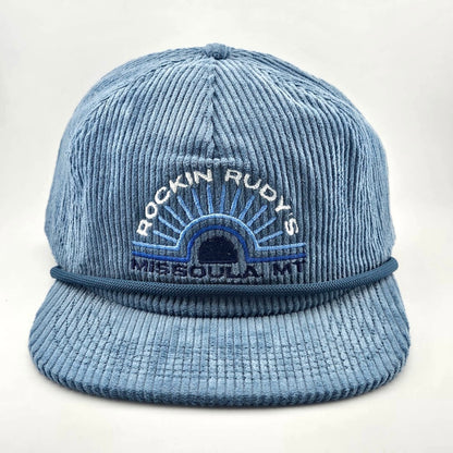 Blue corduroy billed hat with "ROCKIN RUDY'S", radiating from sunburst, "MISSOULA, MONTANA" along horizon bottom.
