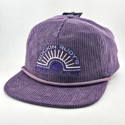 Purple corduroy billed hat with "ROCKIN RUDY'S", radiating from sunburst, "MISSOULA, MONTANA" along horizon bottom.