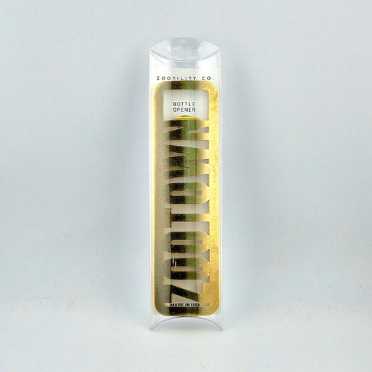 Brass bottle opener reading "ZOOTOWN" in packaging against white background.